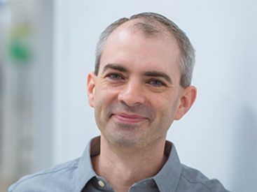 Person with grey hair leaning against white wall in grey shirt