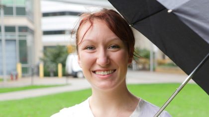 Person with red hair in bun stands outside with umbrella.