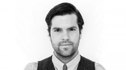 Greyscale headshot of person with beard and black vest, or white background.