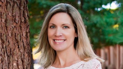 Person with shoulder-length hair standing outdoors next to a tree trunk.