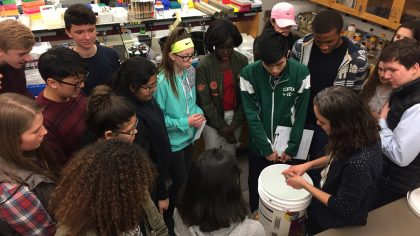 Group of students surrounding an instructor, whose hands are over a white bucket