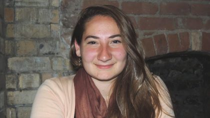 Person with long hair in front of brick wall and fireplace