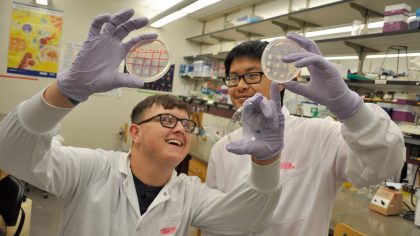 Two male students in lab coats holding up round clear trays/gels