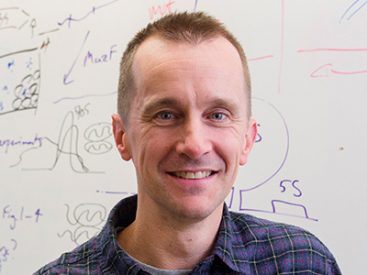 Person in plaid shirt with short hair in front of whiteboard