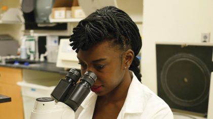 Person with long, dark hair and lab coat stares into microscope.