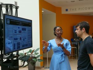 person standing in front of tv screen, talking to a person examining the poster on the tv