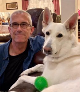 Man sitting on couch next to white dog with pointy ears.